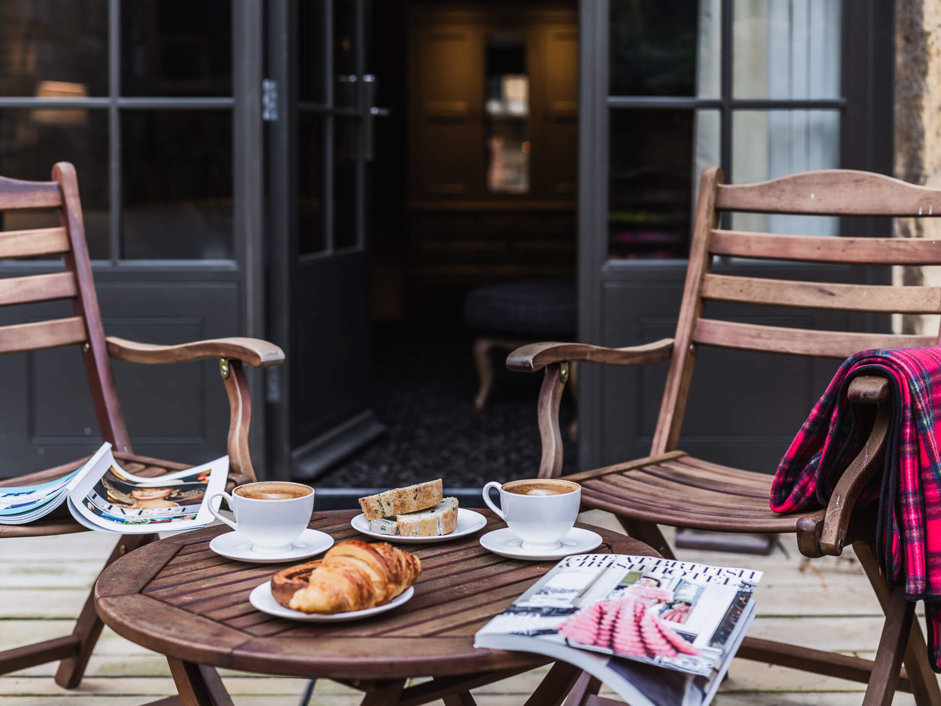 Outdoor seating with coffee, pastries, and magazines on a wooden table.