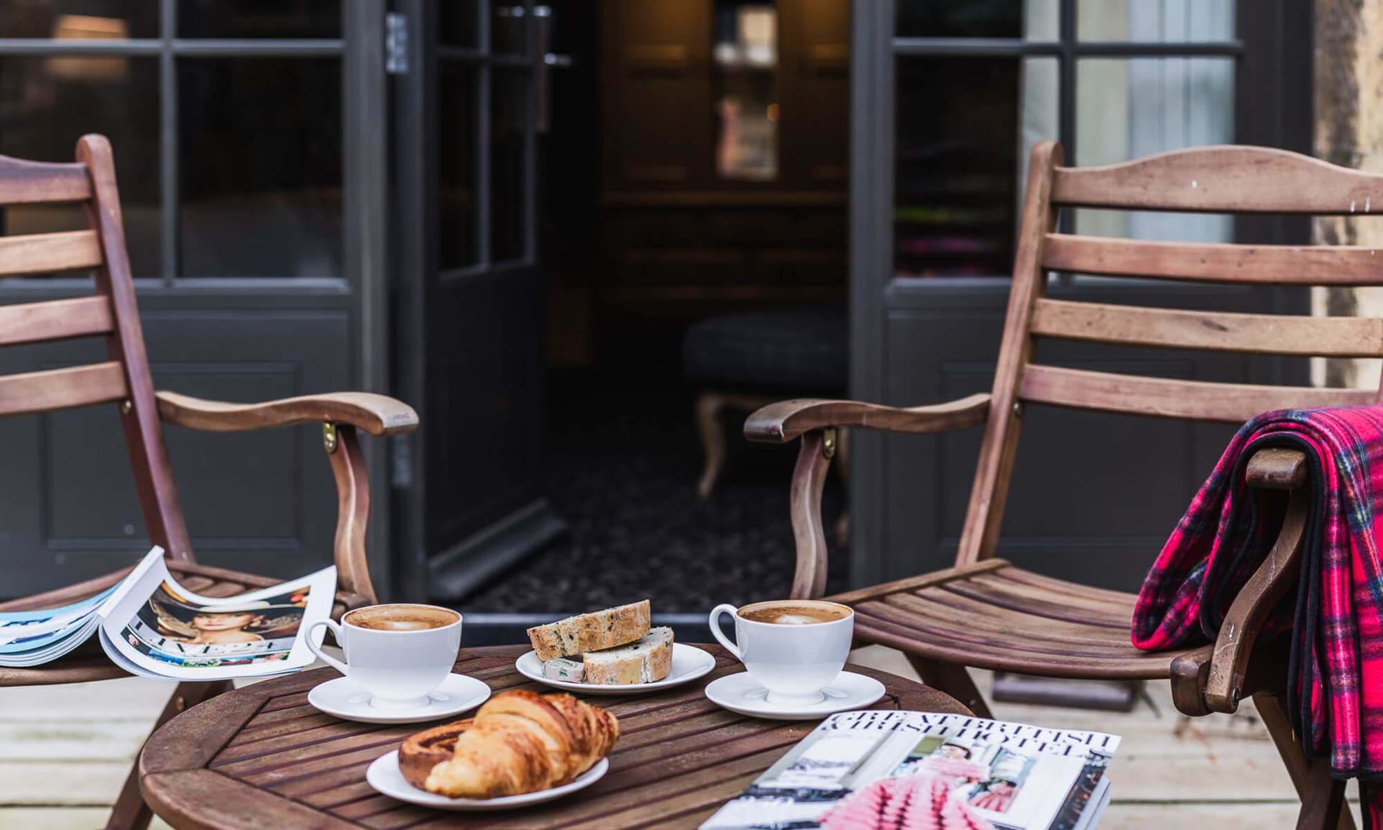 Outdoor seating with coffee, pastries, and magazines on a wooden table.