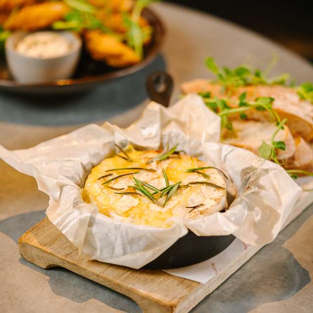 Experience the charm of the best Cotswold pubs with baked cheese and herbs sizzling in a skillet, perfectly paired with sliced bread and a fresh side salad on a rustic wooden board.